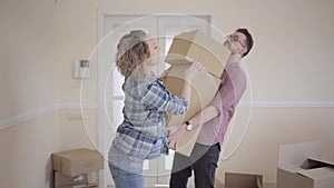 Tall man standing in the room with big box in hands. Woman puts one more box in the top and press on it. Husband nearly