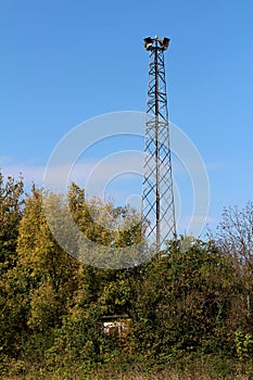 Tall metal structure holding four large public civil defence warning air sirens above small completely overgrown structure and
