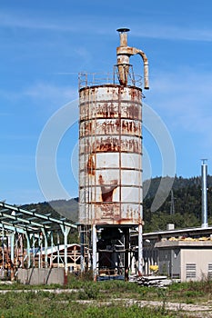 Tall partially rusted metal storage silo surrounded with destroyed buildings at abandoned industrial complex surround with uncut