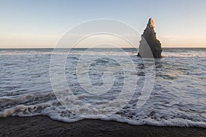 Tall pointy rock on beach shoreline with waves rolling in over sand
