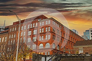 A tall red brick building with some boarded up windows on the street with a blue sky and powerful red and yellow clouds at sunset