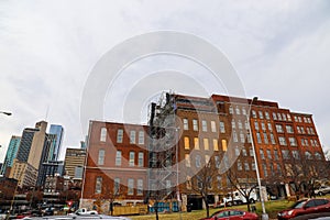 Tall red brick buildings with boarded up and broken windows with bare winter trees in front on the riverfront with cloudy gray sky