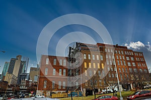 Tall red brick buildings with boarded up and broken windows with bare winter trees in front on the riverfront with cloudy gray sky