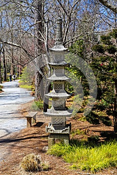 A tall slender stone Japanese lantern in the garden surrounded by brown fallen pine needles and lush green trees and plants