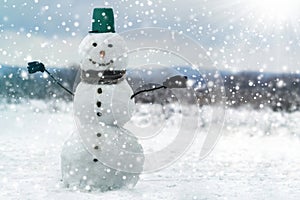 Tall smiling snowman with green bucket hat, scarf and gloves on tree branch hands on white snowy winter landscape, big snowflakes