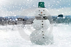 Tall smiling snowman with green bucket hat, scarf and gloves on tree branch hands on white snowy winter landscape, big snowflakes