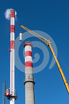 Tall smoking new and renewed old factory chimney with crane boom on blue sky background