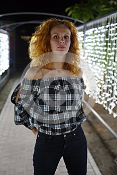 Tall, well-proportioned model-looking girl with red curly hair stands against the backdrop of glowing lights