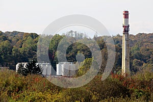 Tall wide concrete red and white rectangle industrial chimney rising high above industrial complex filled with new storage tanks