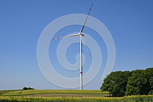 Tall wind turbine with a slender tower and three rotor blades standing on a field in a rural landscape against the blue sky,