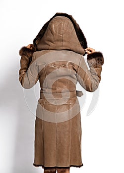 Tall woman model poses with her back to us in luxury brown sheepskin coat with large fluffy fur hood and cuffs on white