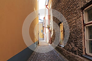Narrow street of a medieval city. The wall is yellow on the left, and an old stone building on the right. Cobblestone road
