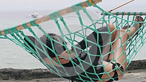 Tan skinned woman lying on a green hammock in front of calm sea.