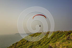 Tandem paragliders on a yellow red parachute fly over the sea and green grass against a clean gray blue sky