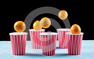 Tangerines fall into multi-colored disposable glasses, close-up on a light blue and black background, copy space