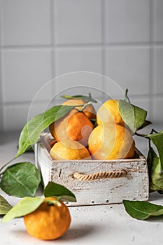 Tangerines (oranges, clementines, citrus fruits) with green leaves in wooden box on white kitchen tile