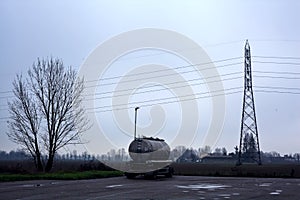 Tank in a parking lot by the edge of a fields with a pylon on a cloudy day