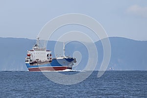 Tanker ship moving in sea full speed ahead on coastal mountains background. Sunny day, calm blue sea, blue sky.
