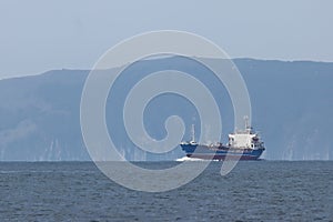 Tanker ship moving in sea full speed ahead on coastal mountains background. Sunny day, calm blue sea, blue sky. Background, design