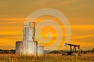 Tanks and pump jack in dry summer field with orange grasses reflecting sunset sky with rolling hills and trees in background