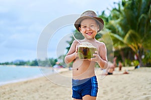 Tanned Asian boy stands on the beach in a hat and drinks coconut