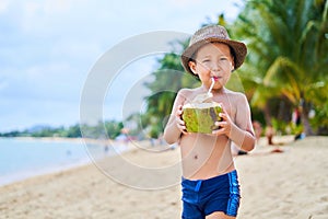 Tanned Asian boy stands on the beach in a hat and drinks coconut