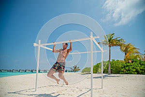Tanned attractive man making pull ups near ocean in swimming wear at tropical beach at island luxury resort
