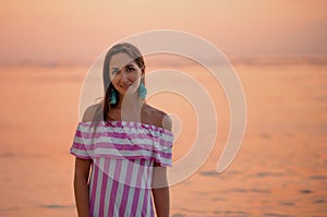 Tanned beautiful woman in dress with white and pink stripes. Close up and copy space. Orange sea or ocean at the sunset. Vacations