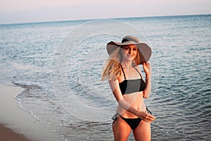 Tanned blonde girl with long hair in a black bikini and a hat smiles and walks along the ocean