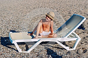 Tanned boy in hat sits on white sunbed on the beach