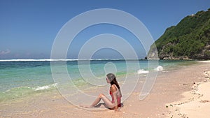 Tanned brunette woman in red swimsuit sitting near water, enjoying coming foam waves on wild tropic beach with blue