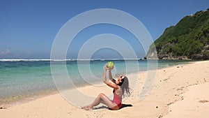 Tanned brunette woman in red swimsuit sitting near water, enjoying coming waves and pour coconut juice over on wild