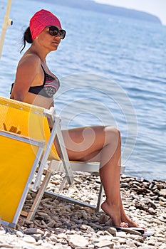 Tanned Caucasian woman sitting under parasol on pebble beach near blue sea water, dressed swimsuit and red kerchief