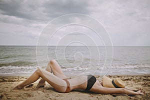 Tanned girl in hat lying on beach. Fashionable young woman covering with straw hat, relaxing on sandy beach near sea. Summer
