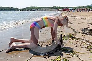 Tanned girl in a rainbow swimsuit plays in the sand on the beach. Sea shore