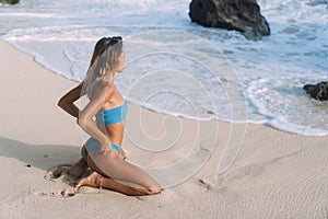 Tanned girl with sporty body in swimsuit sits on sandy beach and looks on ocean and waves