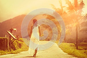 A tanned girl in a white dress walks forward on the road. The view from the back. In the background, a mountain and palm trees.