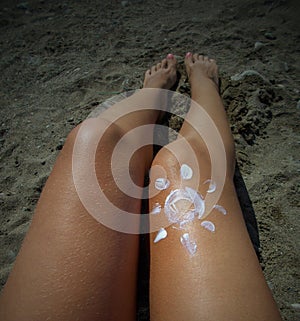 The tanned legs of a young girl with an image of the sun painted with sunscreen. Sandy background. A close-up. Sun cream. Skincare