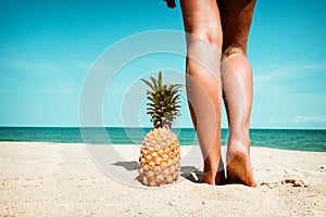 Tanned legs of young woman standing with pineapple at tropical beach in summer