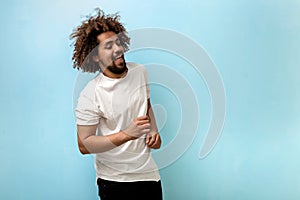 A tanned man with hair in a frizz is broadly smiling while dancing. White T-shirt looks plain and attractive.