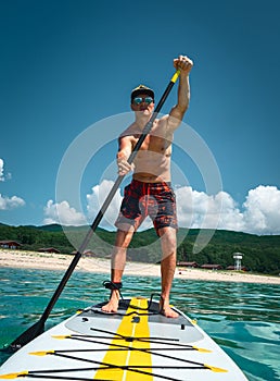 Tanned man stands on a yellow paddle board and paddles. Active sport on the beach in the turquoise sea water