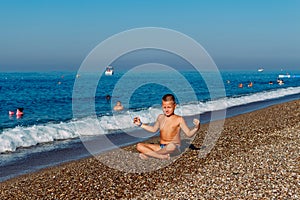 Tanned nine-year-old boy in bathing fits sits back on the pebble shore of the sea