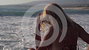 Tanned relaxed woman sitting on coast of Mediterranean sea with foamy waves rolling on beach at sunrise. Carefree happy