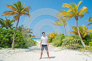 Tanned sexy young man wearing white t-shirt and black shorts standing at tropical sandy beach at island luxury resort