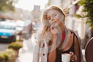 Tanned woman with hair gently waving by wind holding cup of coffee on blur city background. Outdoor portrait of chilling