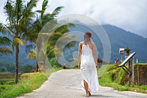 A tanned woman in a white dress walks forward on the road. The view from the back. In the background, a mountain in the fog and