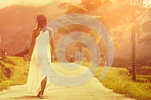 A tanned woman in a white dress walks forward on the road. The view from the back. In the background, a mountain and palm trees.