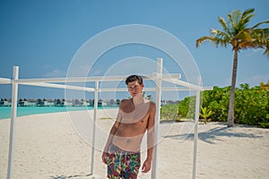 Tanned young sexy man standing near ocean in swimming wear at tropical beach at island luxury resort