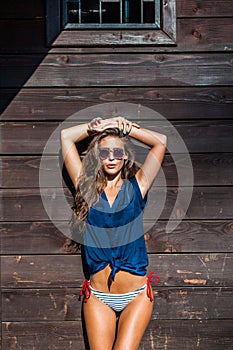 Tanned young woman with sunglasses  in bikini and blue summer shirt by wooden house summer day