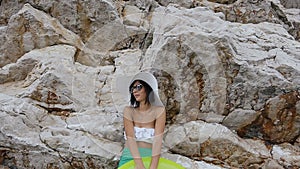 Tanned young woman in white bikini, sunglasses and big hat with mattress near the high rock by the sea. Summer vacation